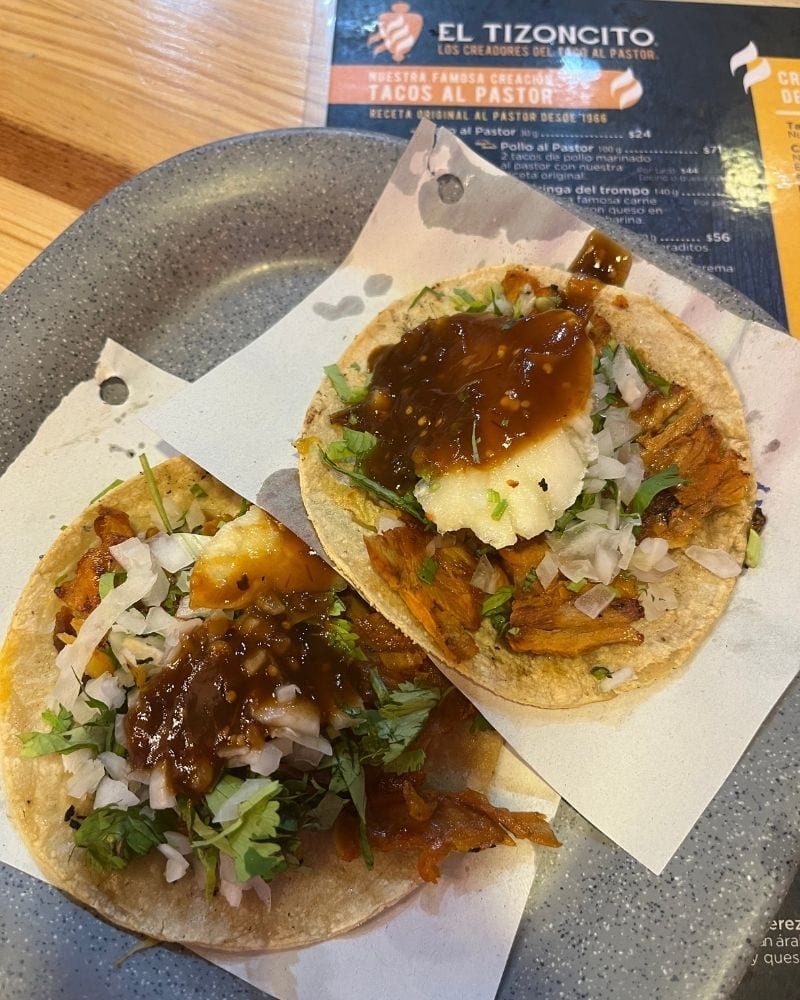 Two tacos al pastor with salsa and pineapple, with the menu behind, at El Tizoncito in Mexico City