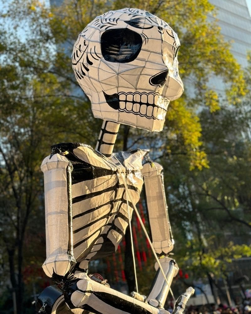 Towering white skeleton figure with a decorated skull against trees and a high-rise in Mexico City for Day of the Dead