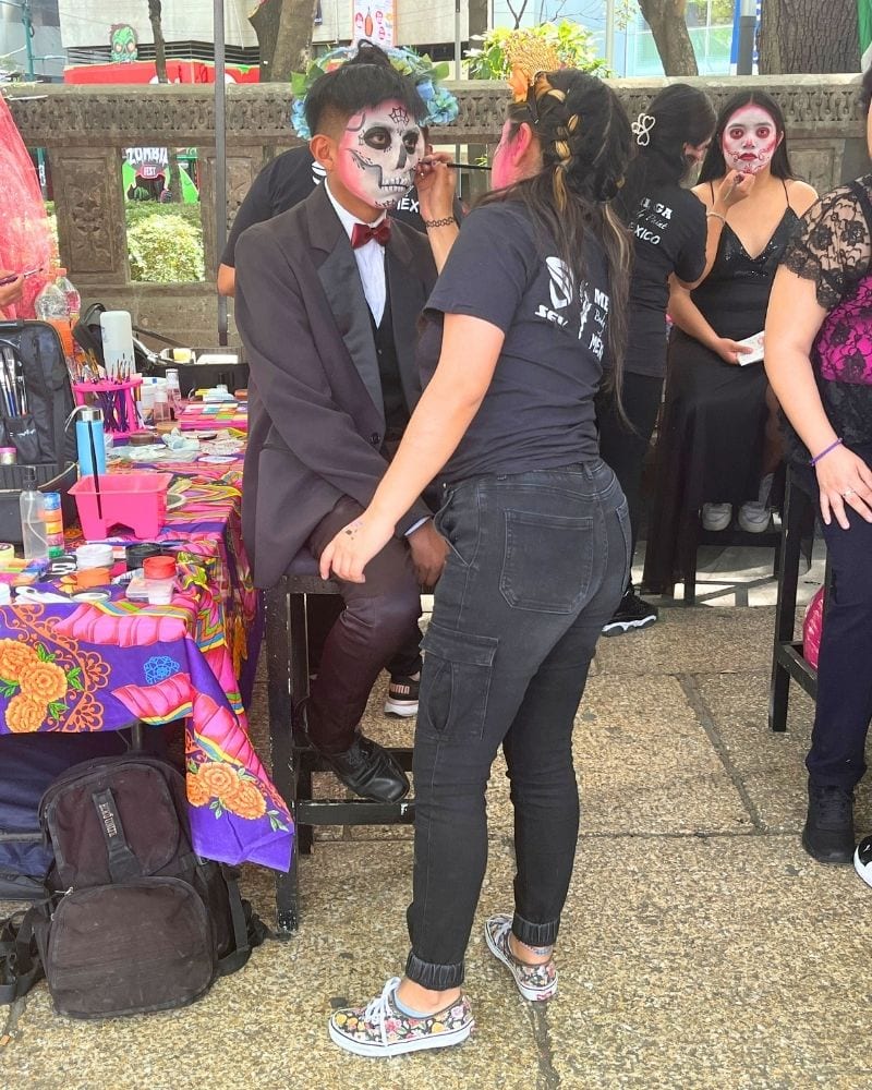 Artist painting a man's face like a calavera at an outdoor face-painting stand in Mexico City for Day of the Dead