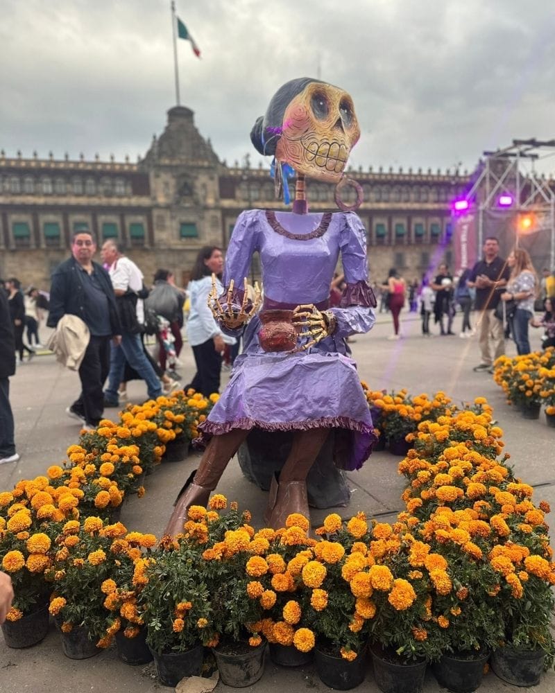 Seated skeleton figure in a lavender dress among marigolds in front of the National Palace in Mexico City for Day of the Dead