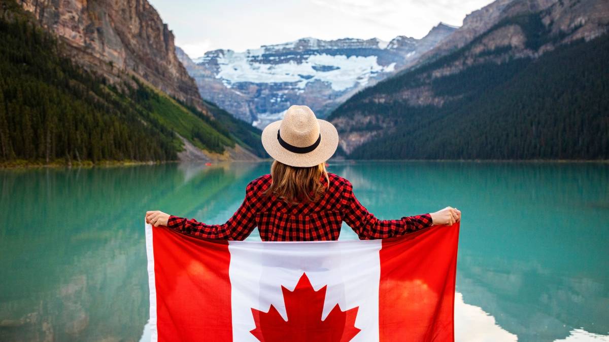 Woman Wearing Red and Black Checkered Dress Shirt and Beige Fedora Hat Holding Canada Flag Looking at Lake