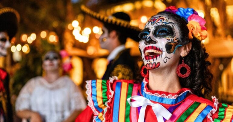 Smiling woman in Catrina face paint with flowers in her hair and a colorful ribboned dress for Day of the Dead