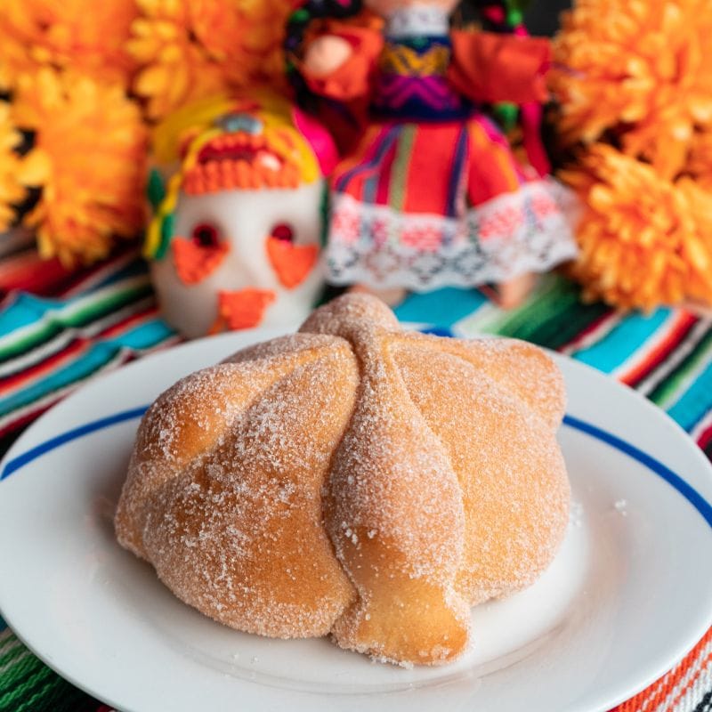Sugar-dusted pan de muerto on a plate in front of marigolds, a sugar skull and a handmade doll for Day of the Dead